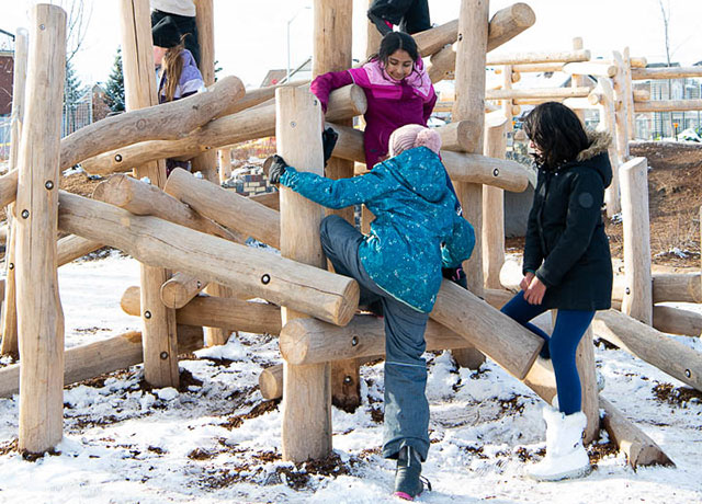 Photo of young children climbing outdoor wooden structure with ropes.