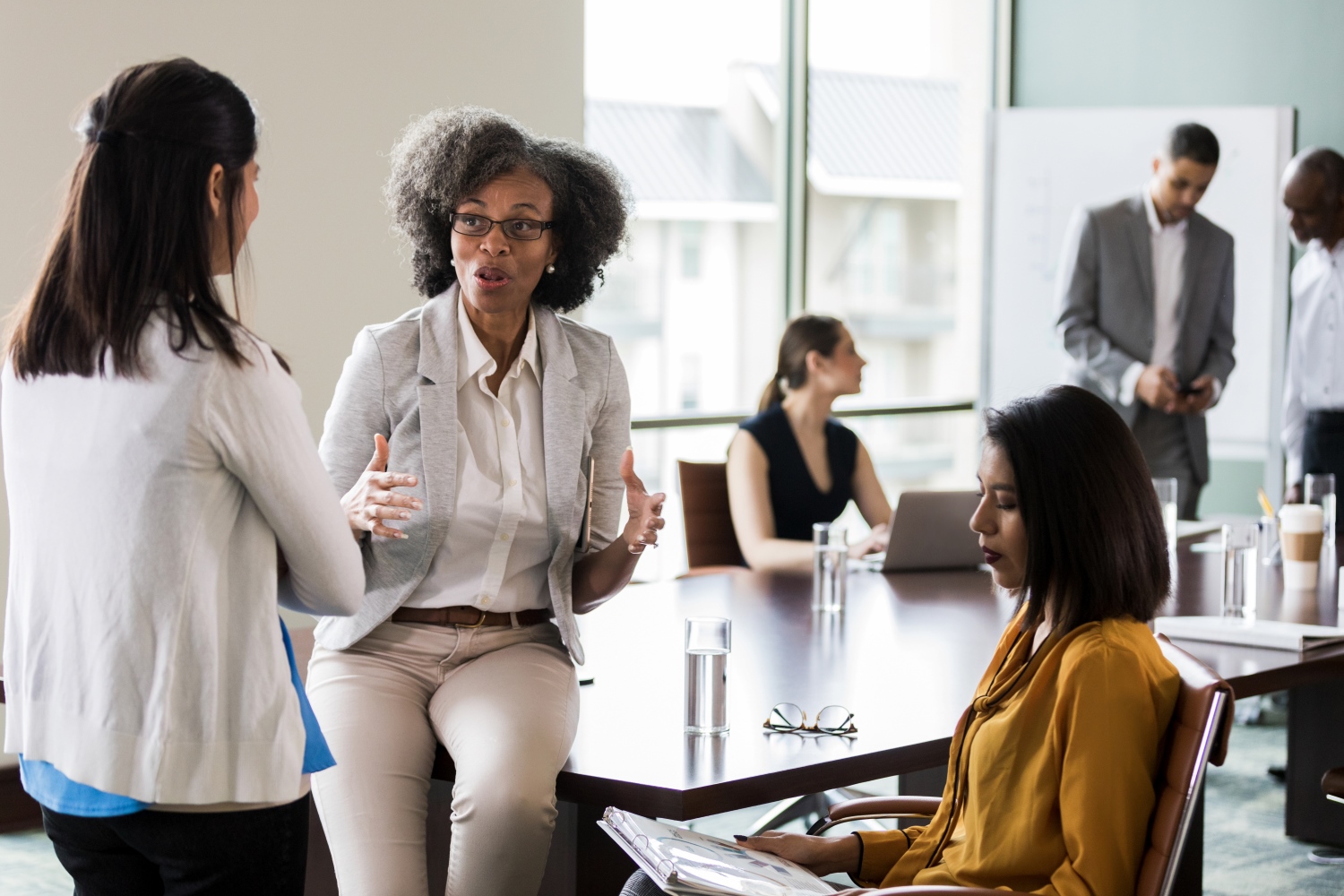 Woman sitting on a table talking to one woman standing with her back to the camera and another sitting looking at a binder. Other professional individuals blurred in background.
