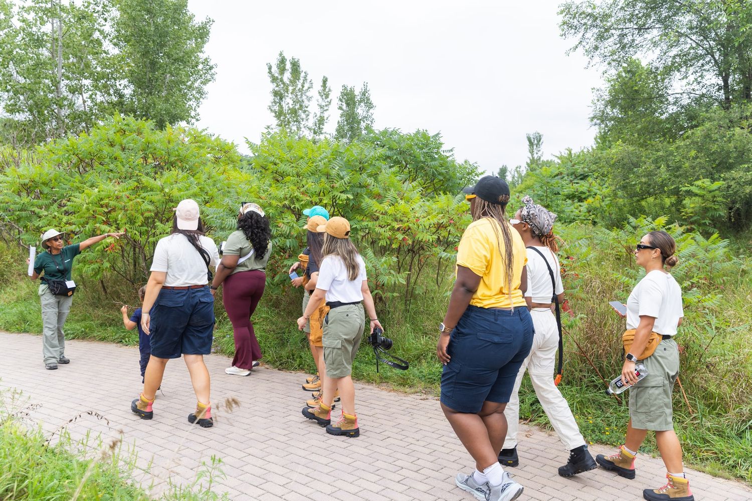 A small group of people walk along a brick path surrounded by green trees, following a leader