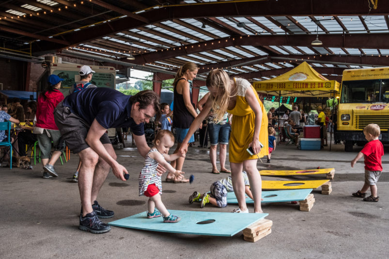A mother and father help a toddler play corn hole outside