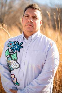 Niigaan Sinclair poses in front of tall prairie grass, wearing a decorative shirt with flowers and a fish.