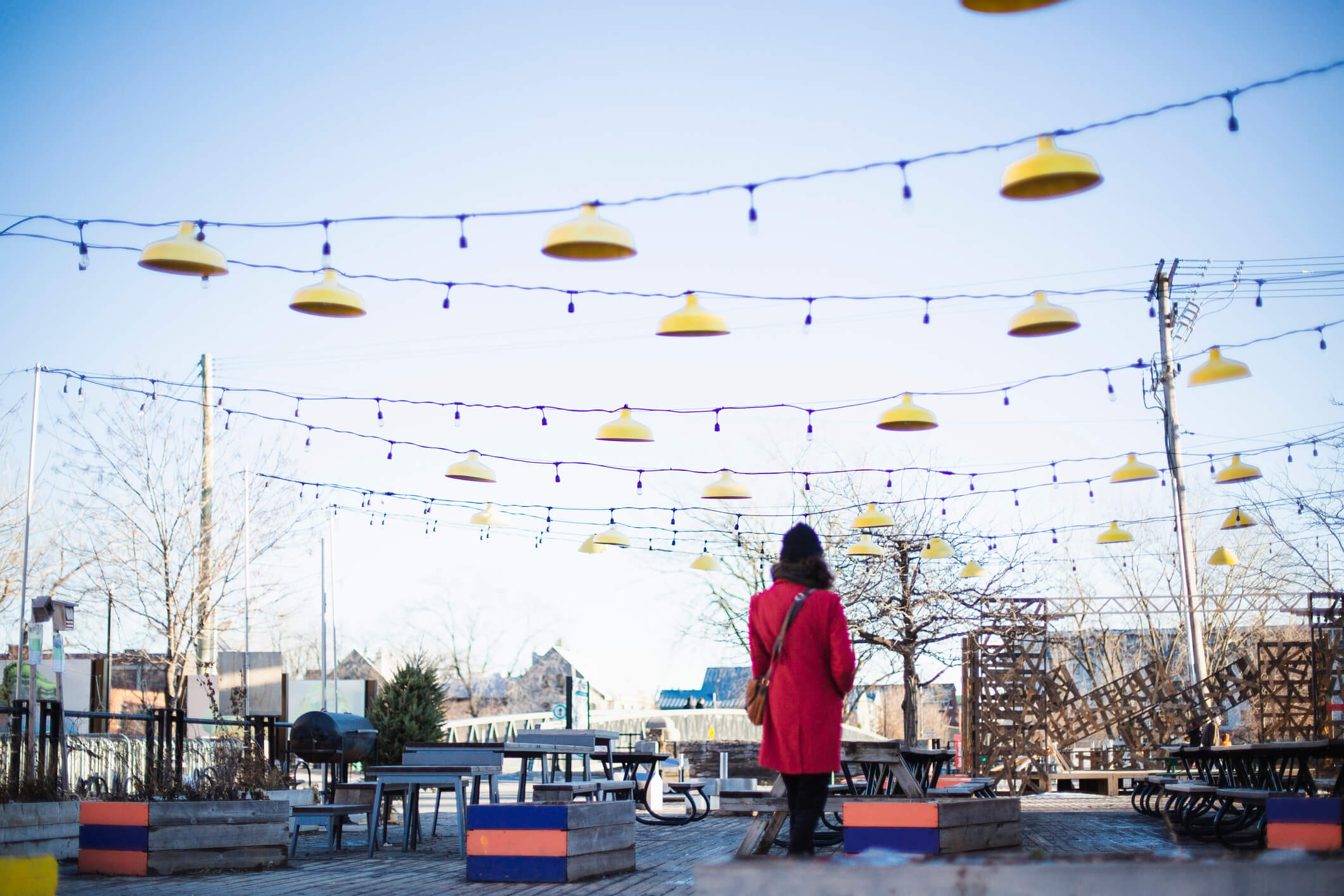 A person in a red coat stands in front of empty picnic tables and string lights